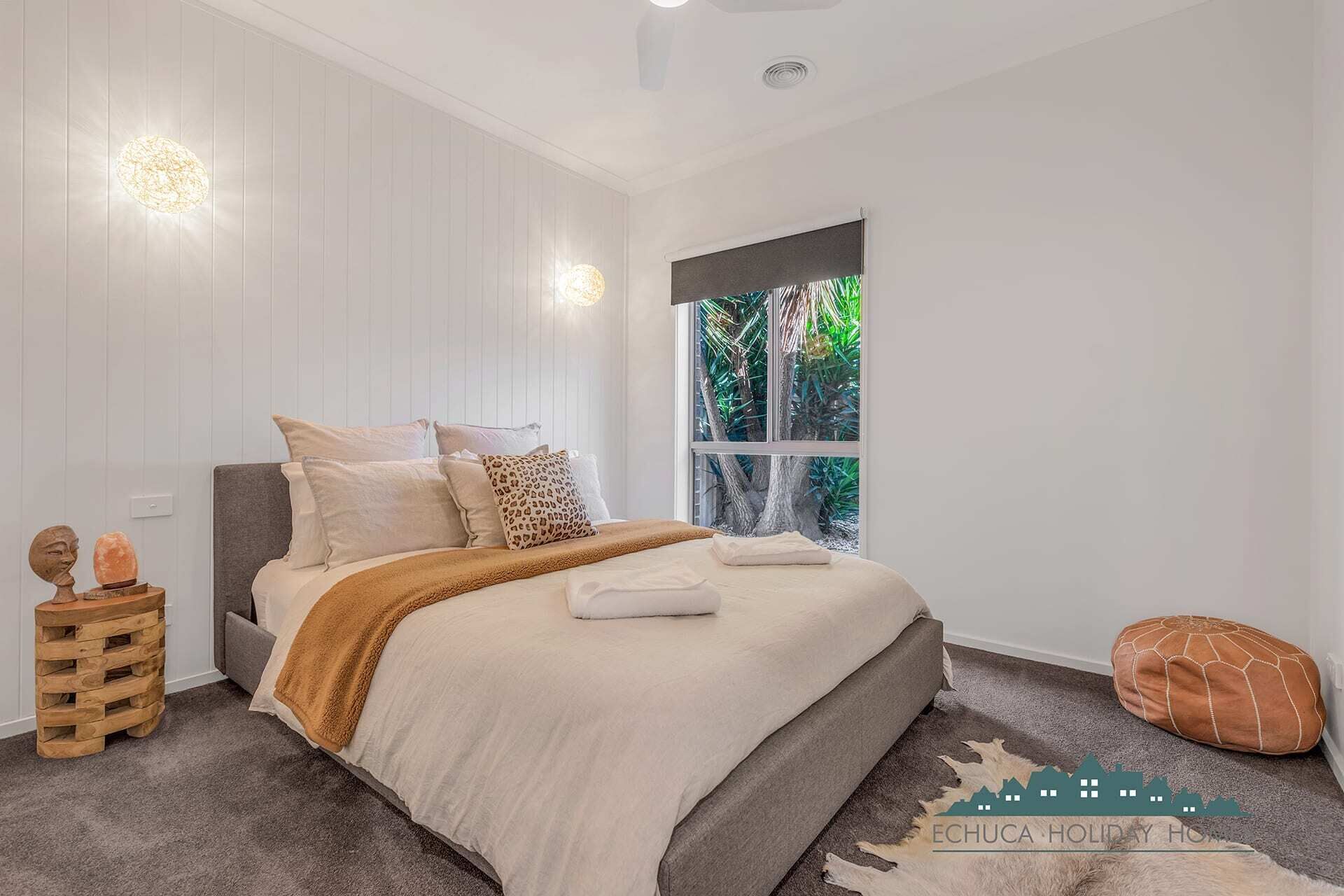 A modern bedroom with a large bed, a window with a view of greenery, and a wooden side table with decorative items.