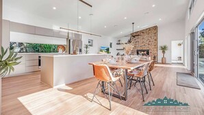 A modern kitchen with a central island, a dining area with a stone fireplace, and a view of the outdoors. - Luxe Haven (Moama)