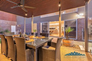 An outdoor dining area with wicker furniture and a glass table, a brick wall, and a modern kitchen visible through glass doors.