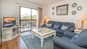 A living room with a sofa, a coffee table, a TV on a stand, and a balcony with a view.