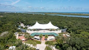 A large, white, tent-like structure with a green roof, surrounded by lush greenery and a body of water in the background.