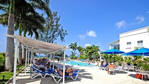 Un espace aménagé au bord de la piscine, avec des chaises longues et des parasols, entouré de palmiers et surplombé d’un ciel bleu azur.