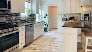 Una cocina moderna con armarios blancos, un refrigerador de acero inoxidable, un horno empotrado y un microondas. Hay plantas en macetas y una cafetera en la encimera.