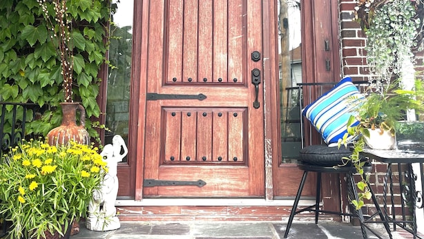 Una puerta de madera con el número '2' encima, rodeada de follaje y una maceta con flores amarillas.