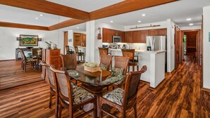 A spacious dining area with a glass-top table surrounded by wooden chairs, a kitchen with wooden cabinets and stainless steel appliances, and a wooden floor throughout the space.