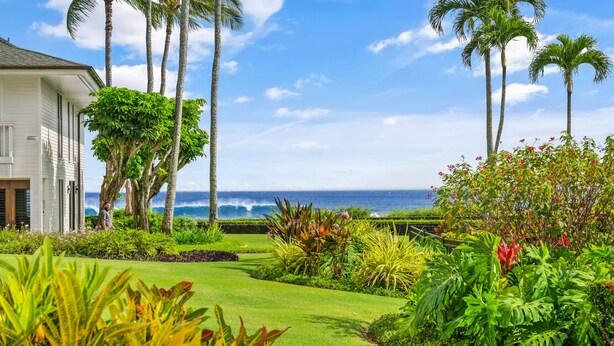 A lush garden with palm trees and tropical plants, leading to a white house with a view of the ocean.