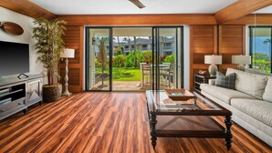 A modern living room with a wooden floor, a ceiling fan, a flat-screen TV, a sofa, a coffee table, and a sliding glass door leading to a garden.