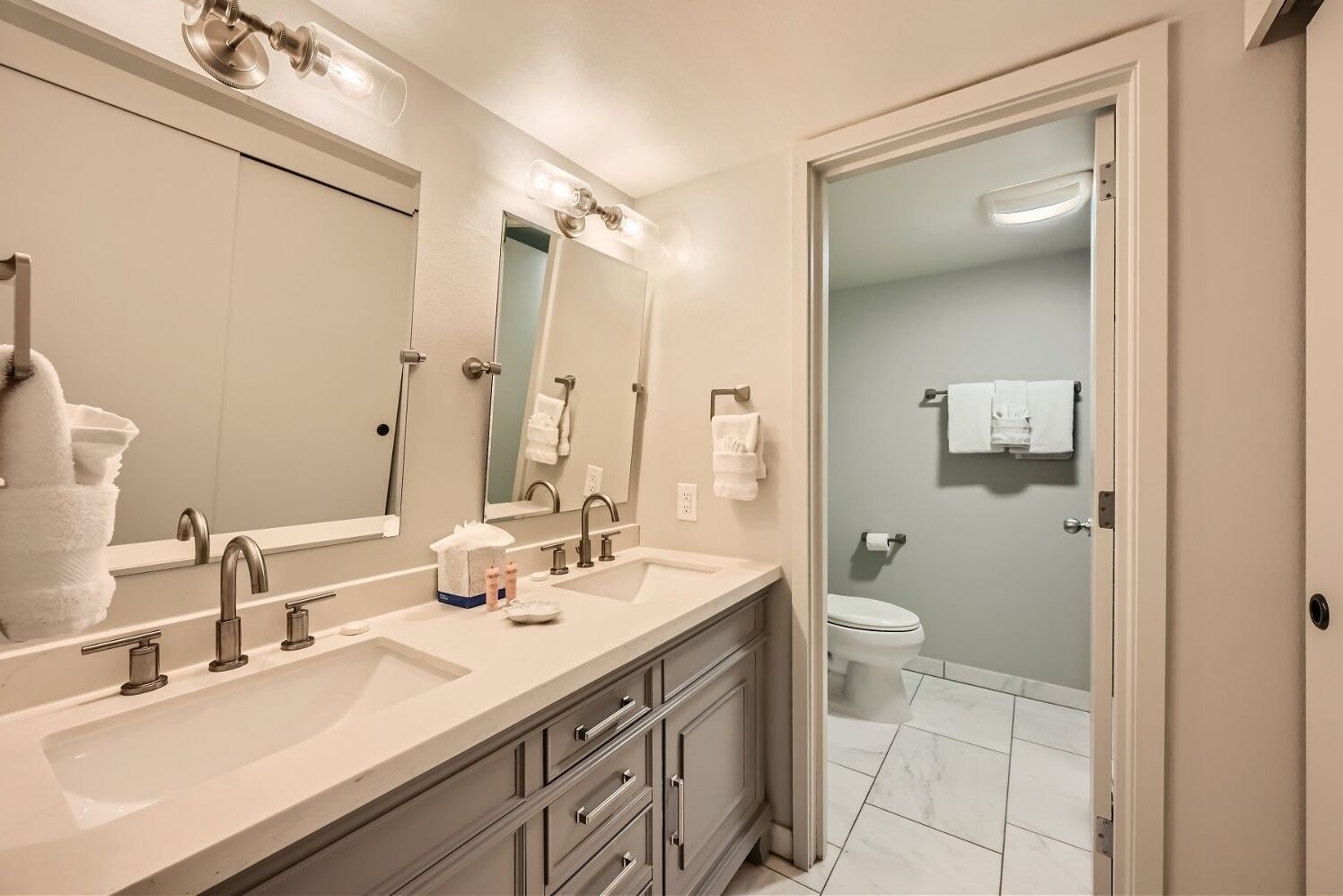 A modern bathroom with a double sink, large mirrors, and a toilet visible through a glass door.
