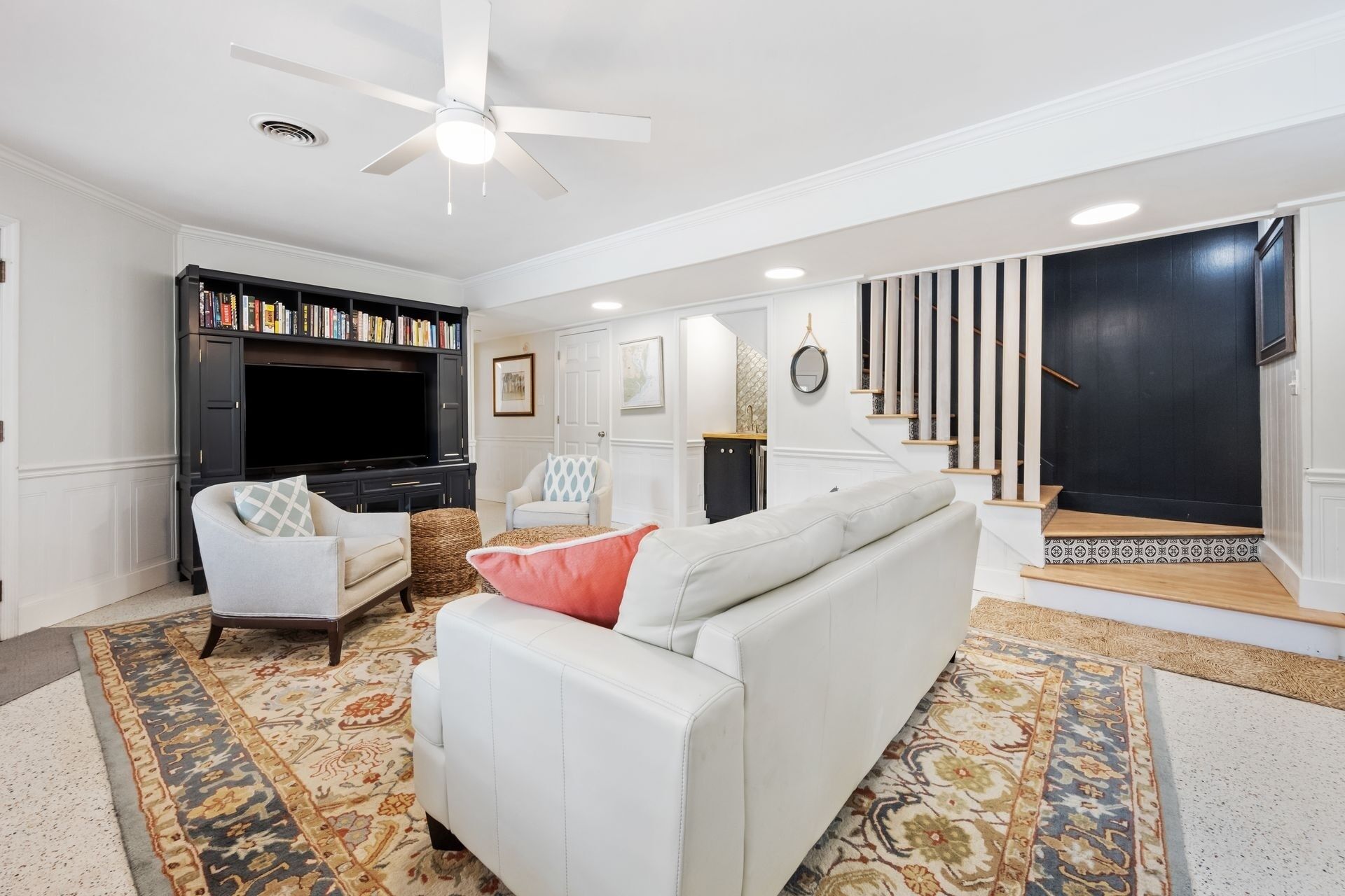 A living room with a white sofa, a bookshelf, a flat-screen TV, and a staircase leading to a dark room.