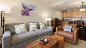 A modern living room with a grey sofa, a brown leather chair, and a wooden coffee table. A painting of a dog is on the wall, and there is a kitchen area in the background. - Base Camp 304 1 Bedroom Condo by RedAwning (Granby)