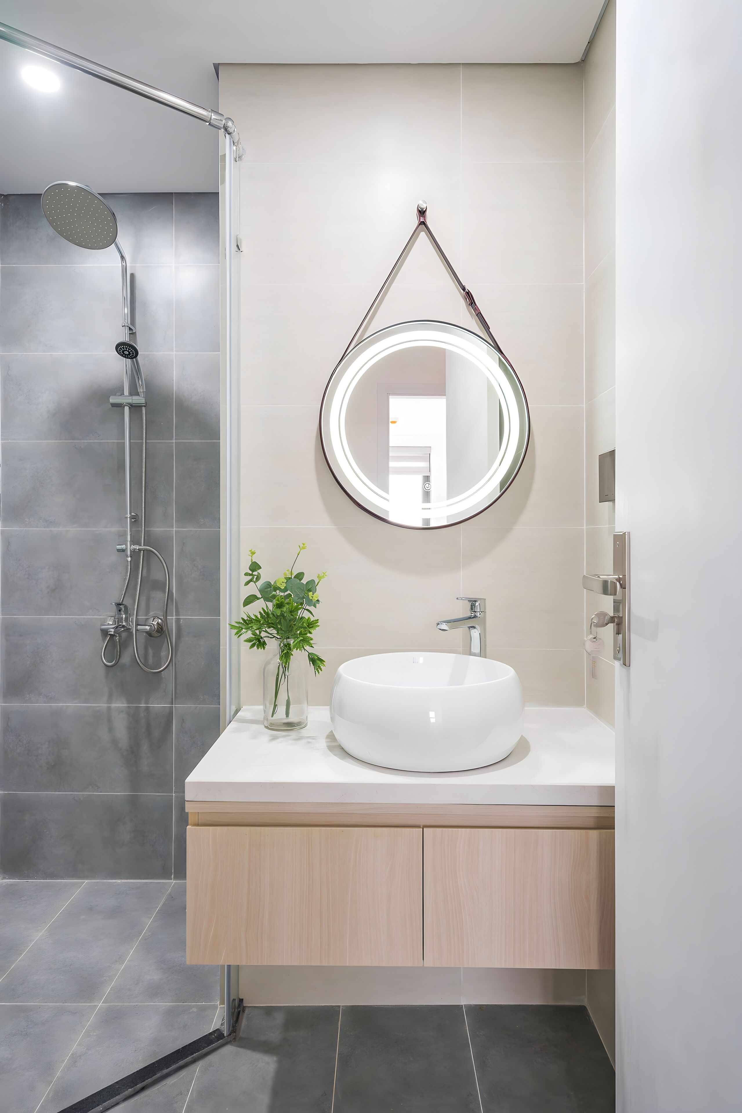 A modern bathroom with a glass-enclosed shower, a round mirror, a white sink, and a wooden cabinet.