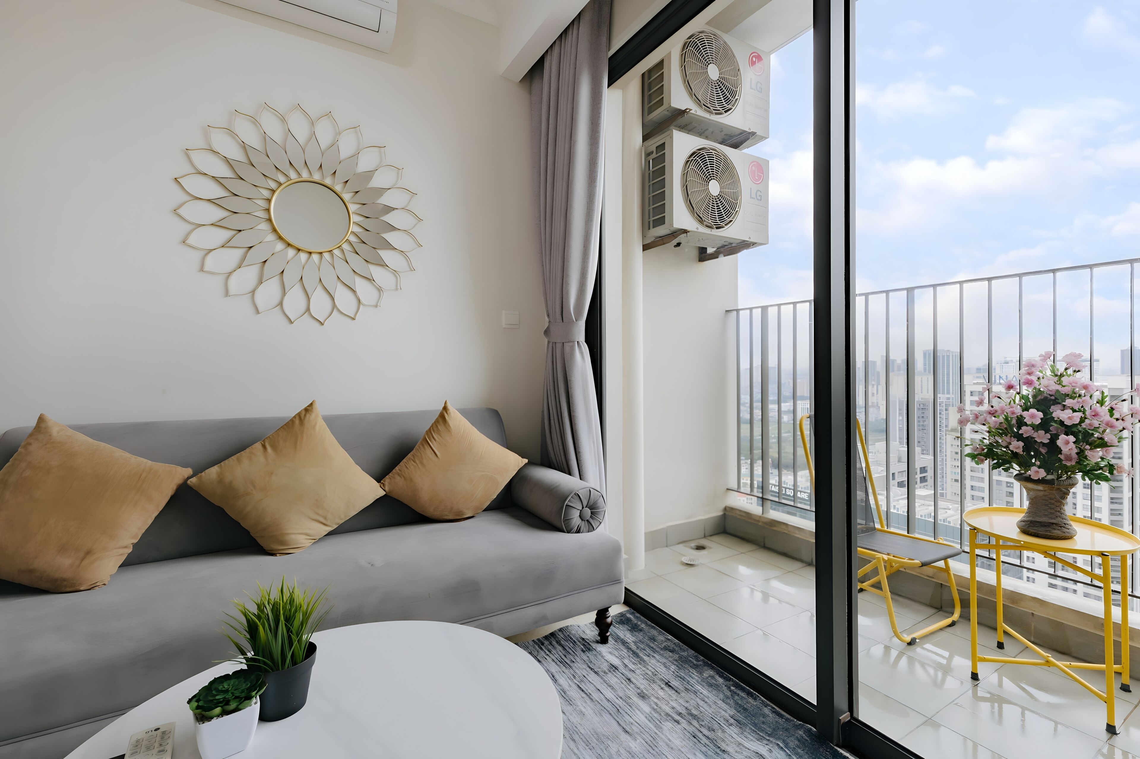 A modern living room with a grey sofa, two beige cushions, a round coffee table, a potted plant, and a yellow chair by a large window with a city view.