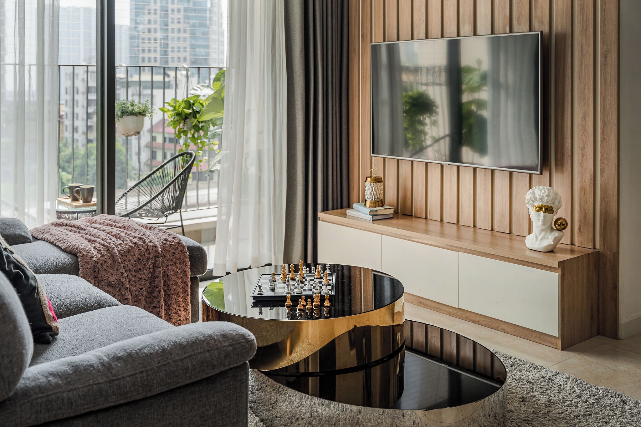 A modern living room with a sofa, a coffee table with a chess set, a TV mounted on the wall, and a balcony with plants.
