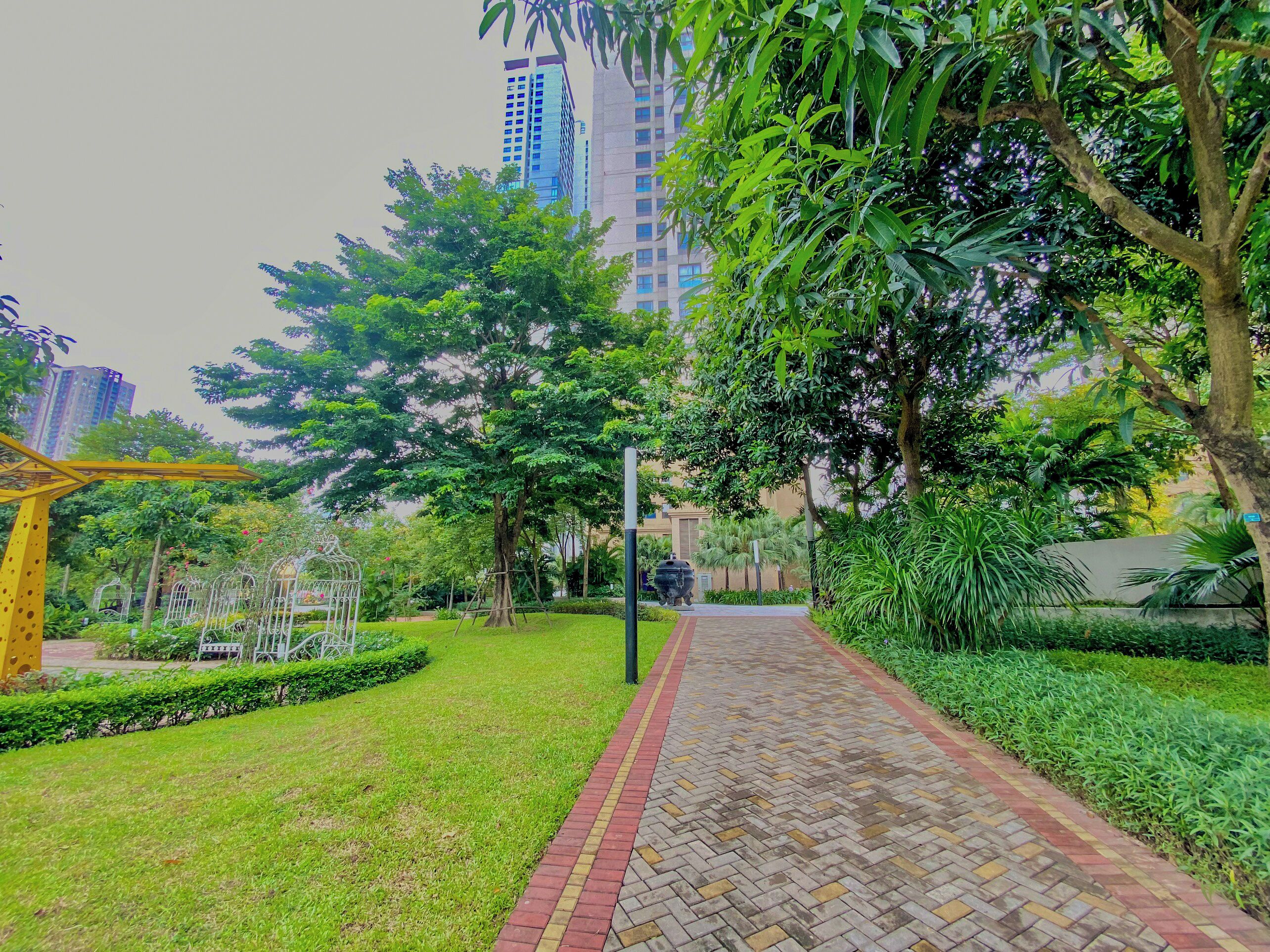 A pathway lined with greenery and a yellow structure, likely a playground, in a residential area with tall buildings in the background.