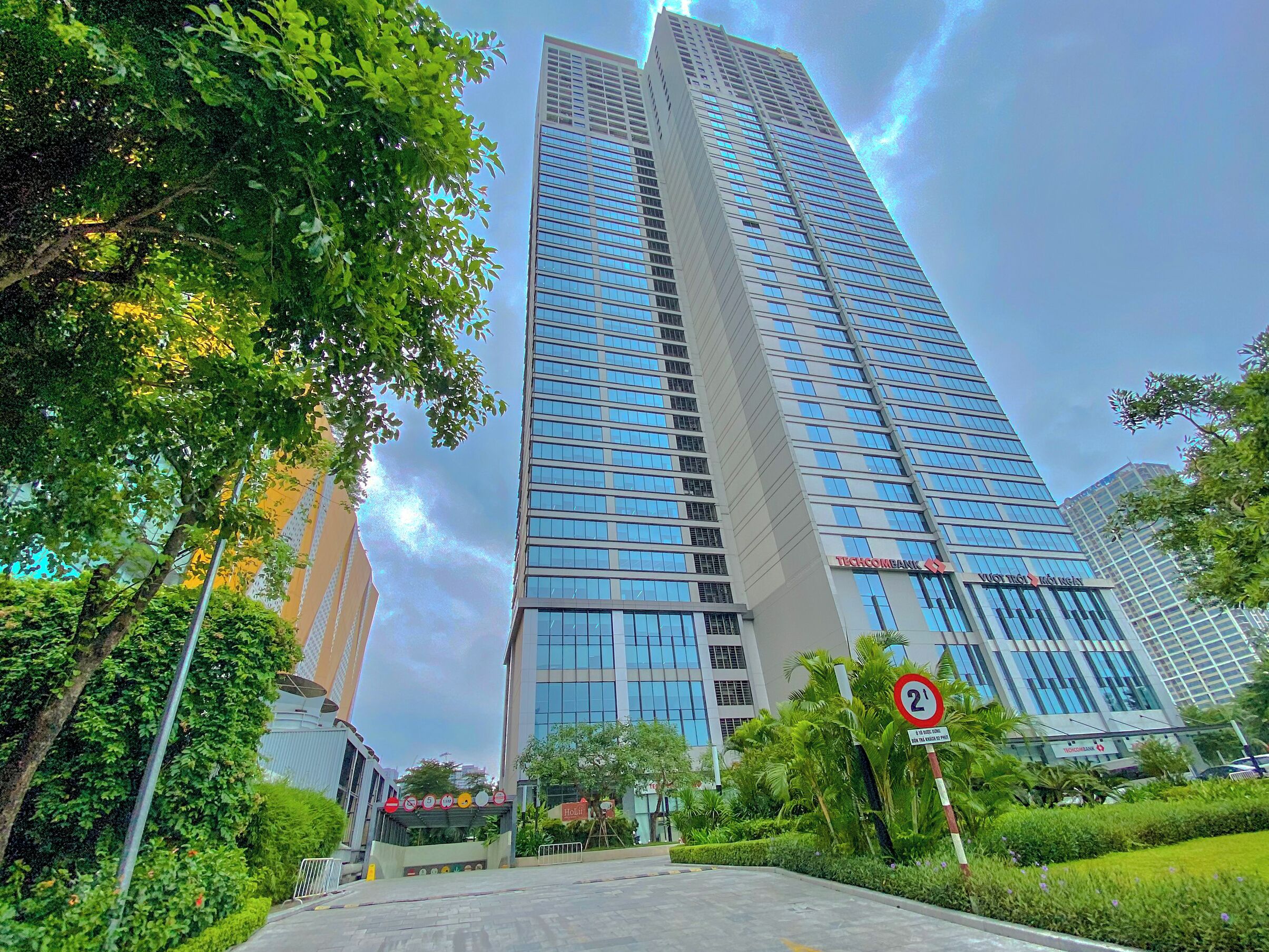 A tall, modern skyscraper with a glass facade, surrounded by lush greenery and palm trees.