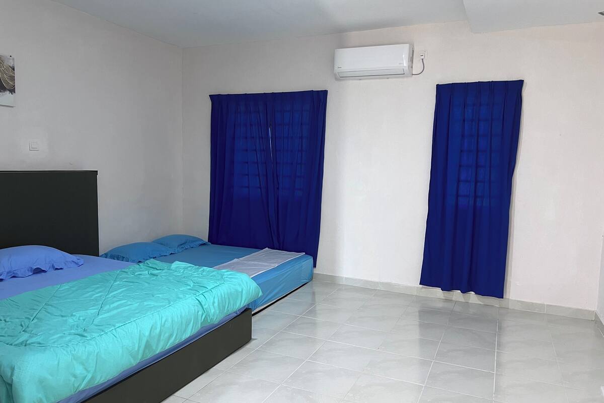 A room with two beds, a window with blue curtains, an air conditioning unit, and a tiled floor.
