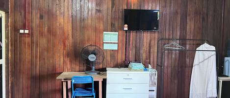 A room with wooden panel walls, a desk with drawers, a chair, a standing fan, a TV mounted on the wall, and a clothes drying rack with a towel.