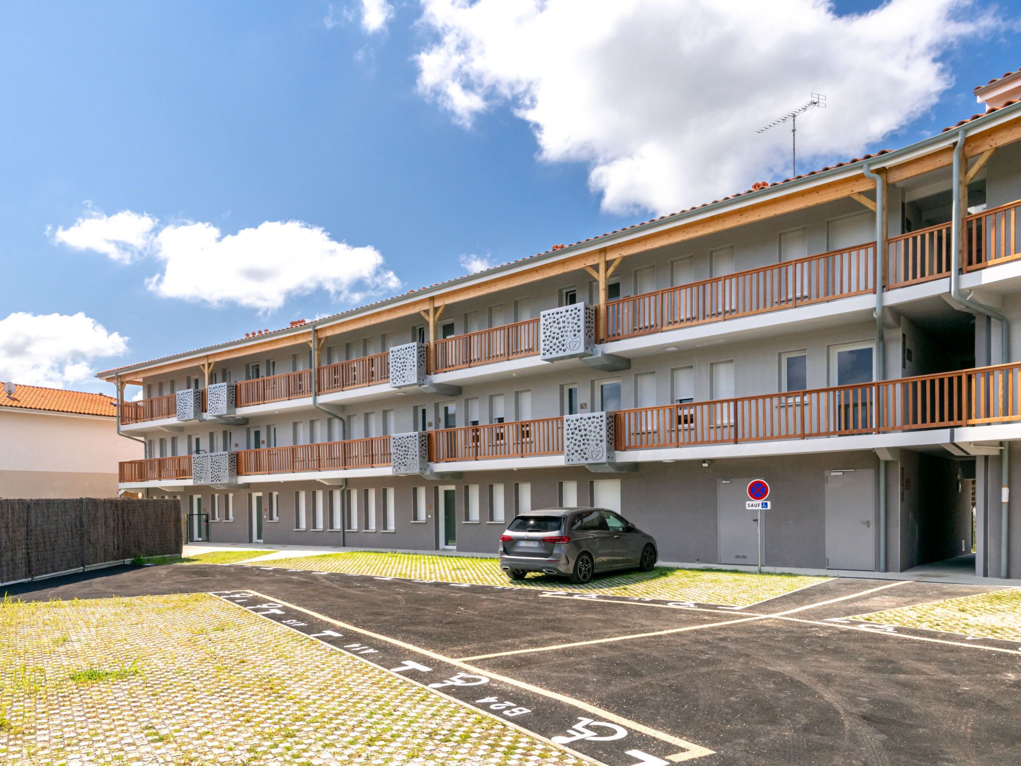 A multi-story building with balconies, a parking area with a car, and a clear sky.