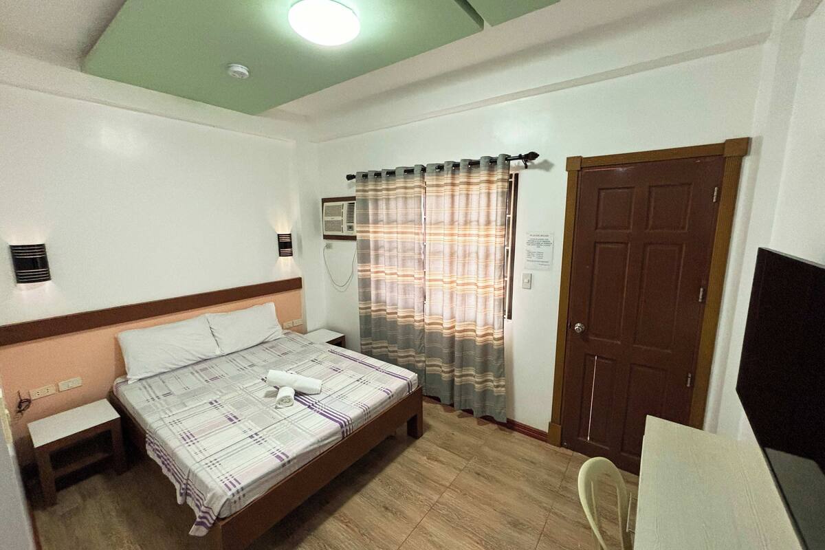 Une chambre d’hôtel comprenant un lit, une table de chevet, une lampe, un appareil de climatisation, des rideaux et une porte.