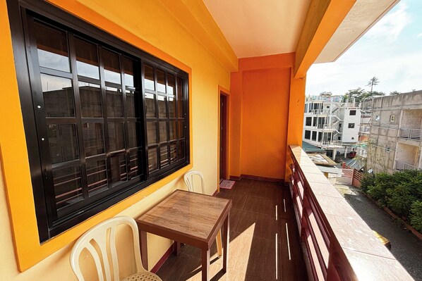 Un balcón con mesa y sillas de madera, una ventana con marcos negros y vista a edificios al fondo.