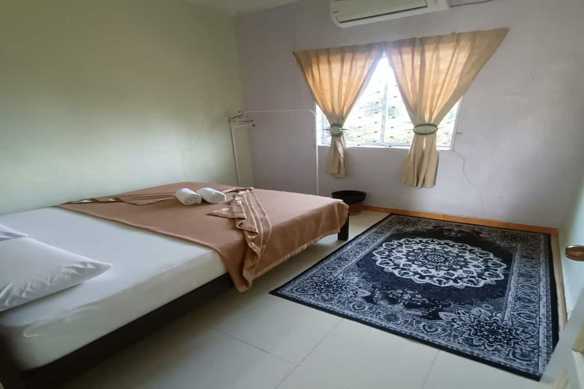 A small, simple room with a bed, a window with curtains, an air conditioning unit, and a patterned rug.
