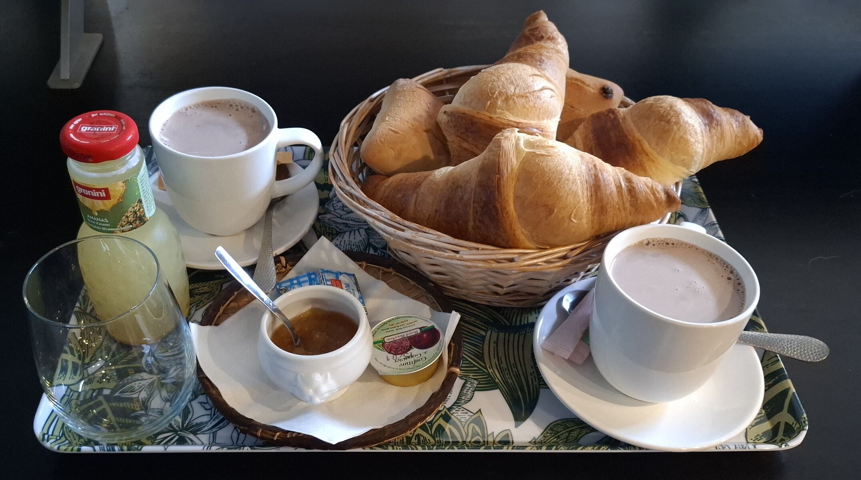 A tray with croissants, two cups of coffee, a jar of granini, and a bowl of jam.