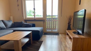 A modern living room with a sofa, a TV, a wooden coffee table, and a view through a sliding glass door. - Apartment 'Wohner' with Private Garden, Balcony and Wi-Fi (Petersaurach)