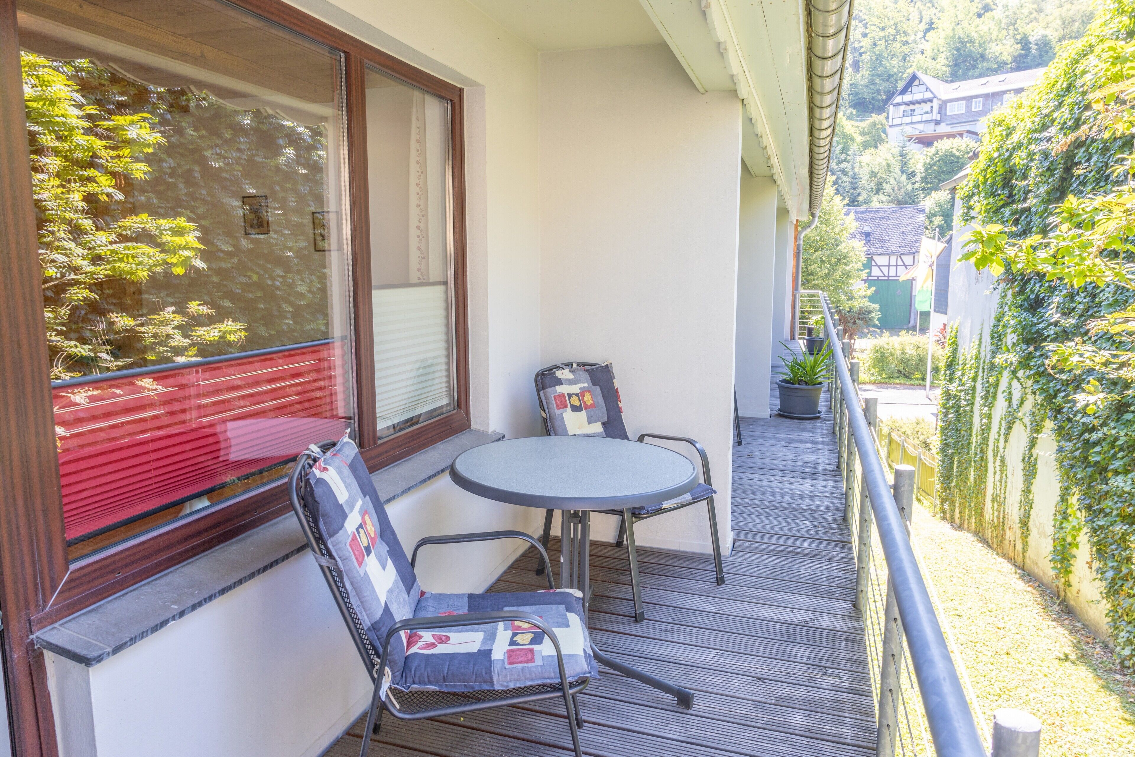 A balcony with a table and two chairs, a window with blinds, and a view of a house and trees.