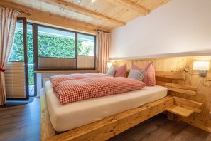Een houten bed met een geruit plaid, een raam met jaloezieën en een lamp aan de muur.