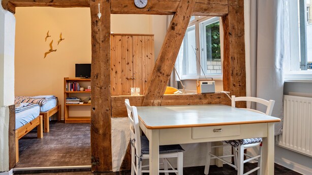 A room with a wooden ceiling structure, a white table with chairs, a bed with a blue bedspread, a bookshelf, and a window with a view of greenery. - Studio Apartment 'Ferienwohnung Stralsund' with Wi-Fi (Stralsund)