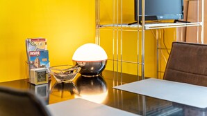 A hotel room with a yellow wall, a desk with a lamp, a chair, a clock, and a television on a shelf. - Studio Apartment 'Ferienwohnung Rügen' with Wi-Fi (Stralsund)