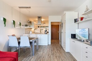 Une cuisine moderne avec des meubles blancs, un coin repas avec deux chaises, un canapé rouge et un téléviseur posé sur un meuble blanc.
