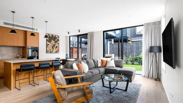 Ein modernes Wohnzimmer mit einer Couch, einem Couchtisch und Barhocker. Es gibt eine Küchenzeile mit Holzausziehtischen und einen Esstisch. Das Zimmer verfügt über große Fenster und einen Blick ins Freie.