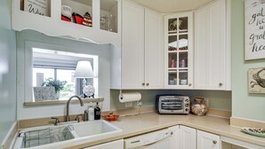 Eine Küche mit weißen Schränken, einer Spüle, einem Fenster mit Ausblick, einem Toaster und einem Einmachglas mit Meeresschnecken.