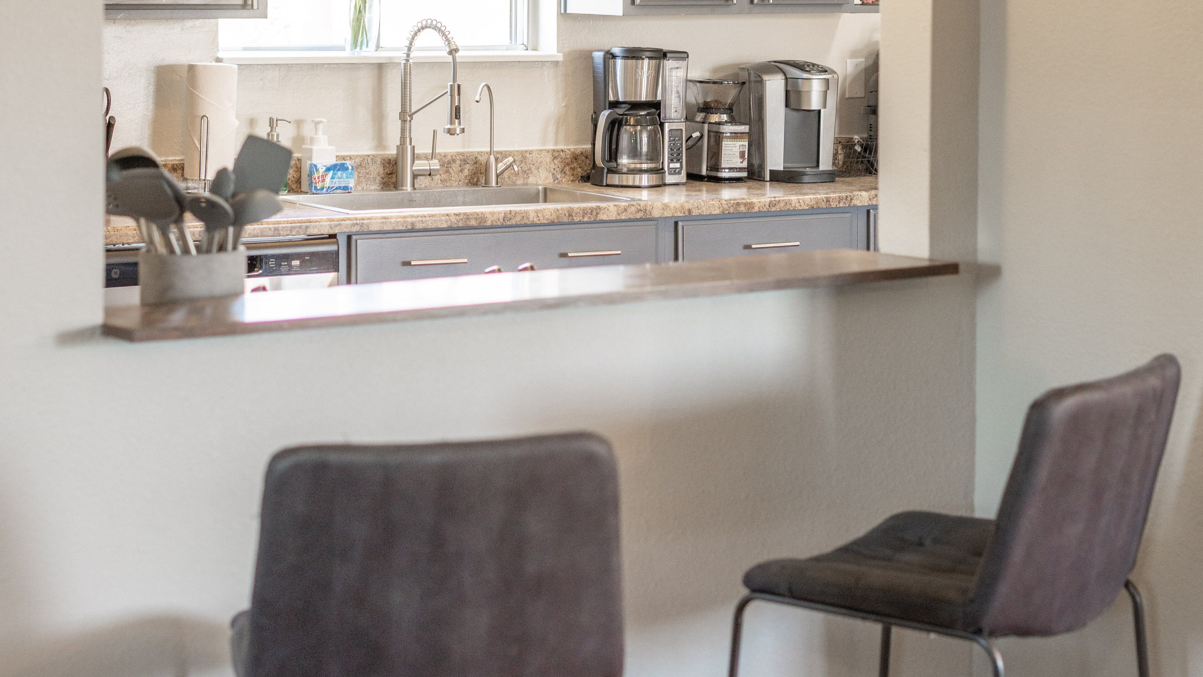 A kitchen with a stainless steel sink, a coffee maker, and a microwave. There are two chairs in the foreground.