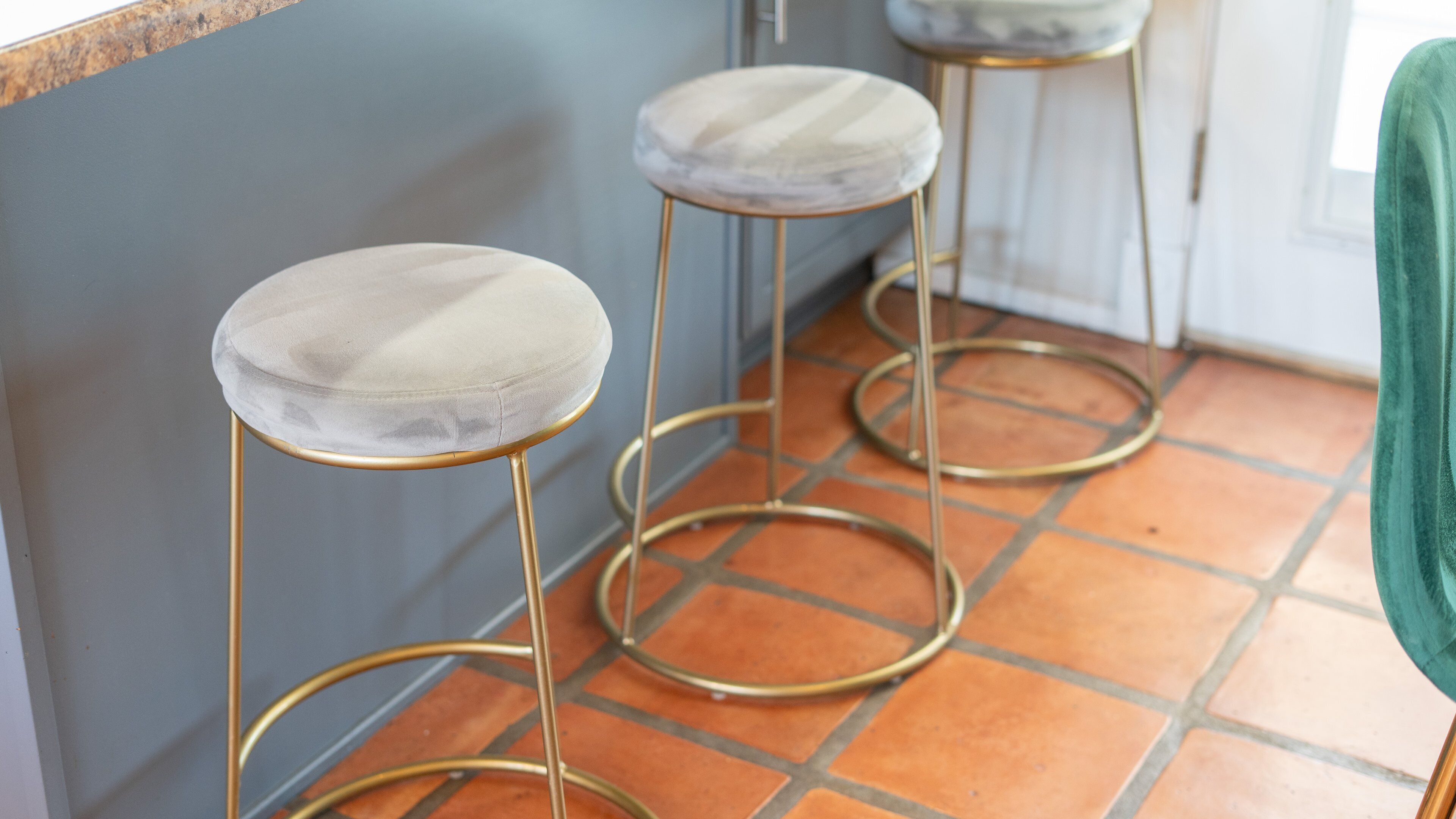 Three bar stools with marble tops and gold legs, positioned against a blue wall with a tiled floor.