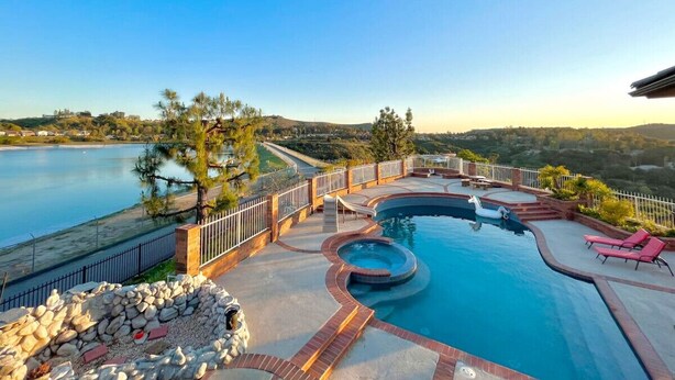 A swimming pool with a hot tub, surrounded by a deck with lounge chairs, a stone wall, and a view of a lake and hills. - Stunning Panoramic Waterfront Pool Home Escape (Anaheim)