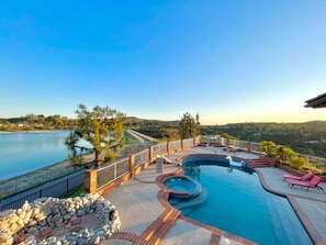 Una piscina con jacuzzi, rodeada por una terraza con sillas de descanso, un muro de piedra y vistas a un lago y colinas.