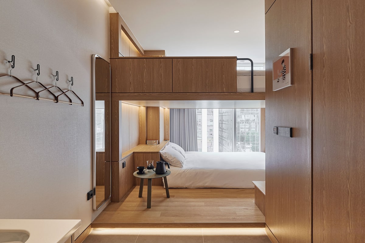 A compact hotel room with a bunk bed, a small table with a kettle, and a view of the city through large windows.