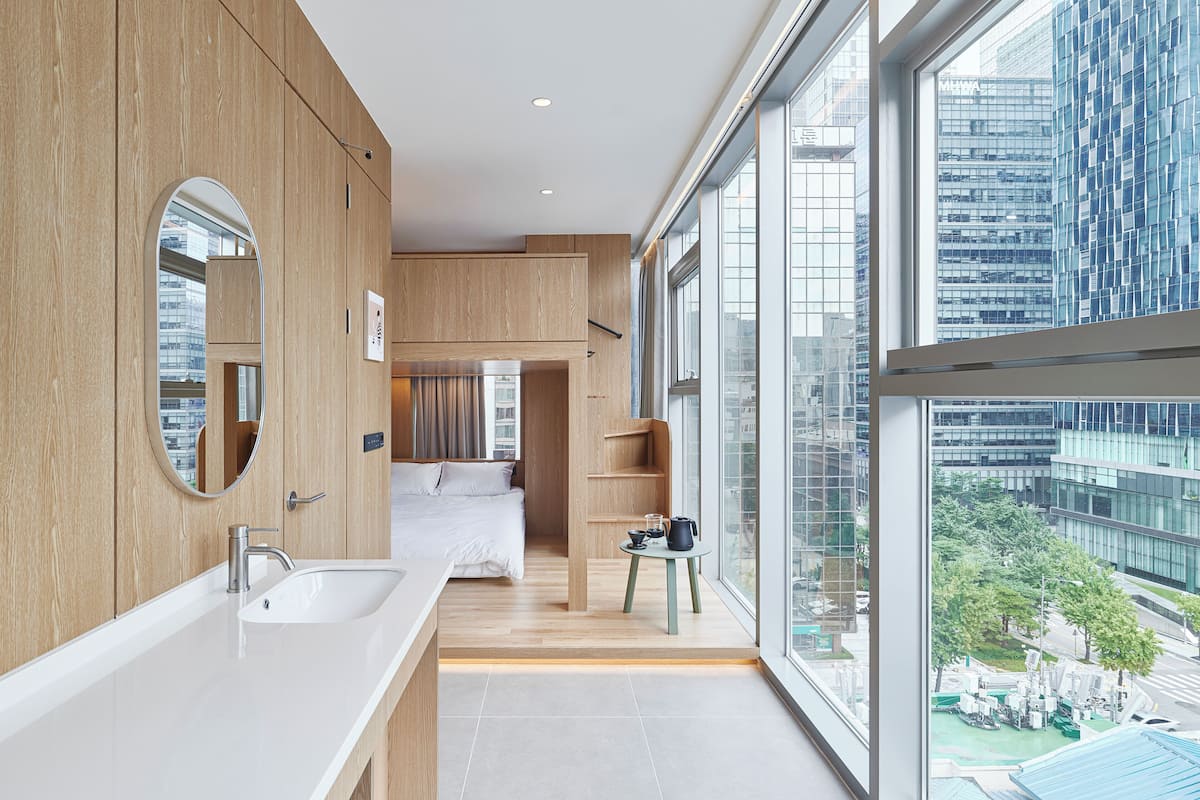 A modern hotel room with a large window, a wooden vanity, and a bed with a view of the city.