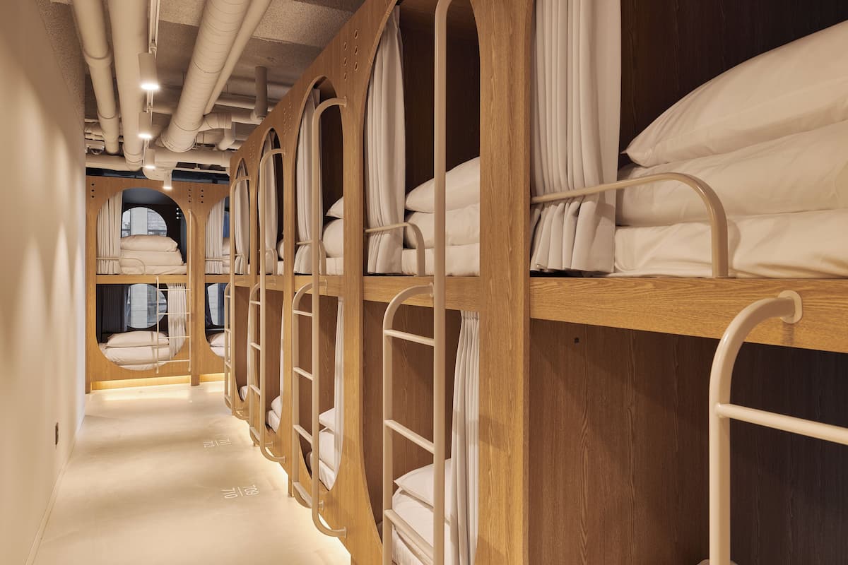 A row of bunk beds with wooden frames and white bedding, arranged in a narrow corridor with a wooden floor and ceiling.