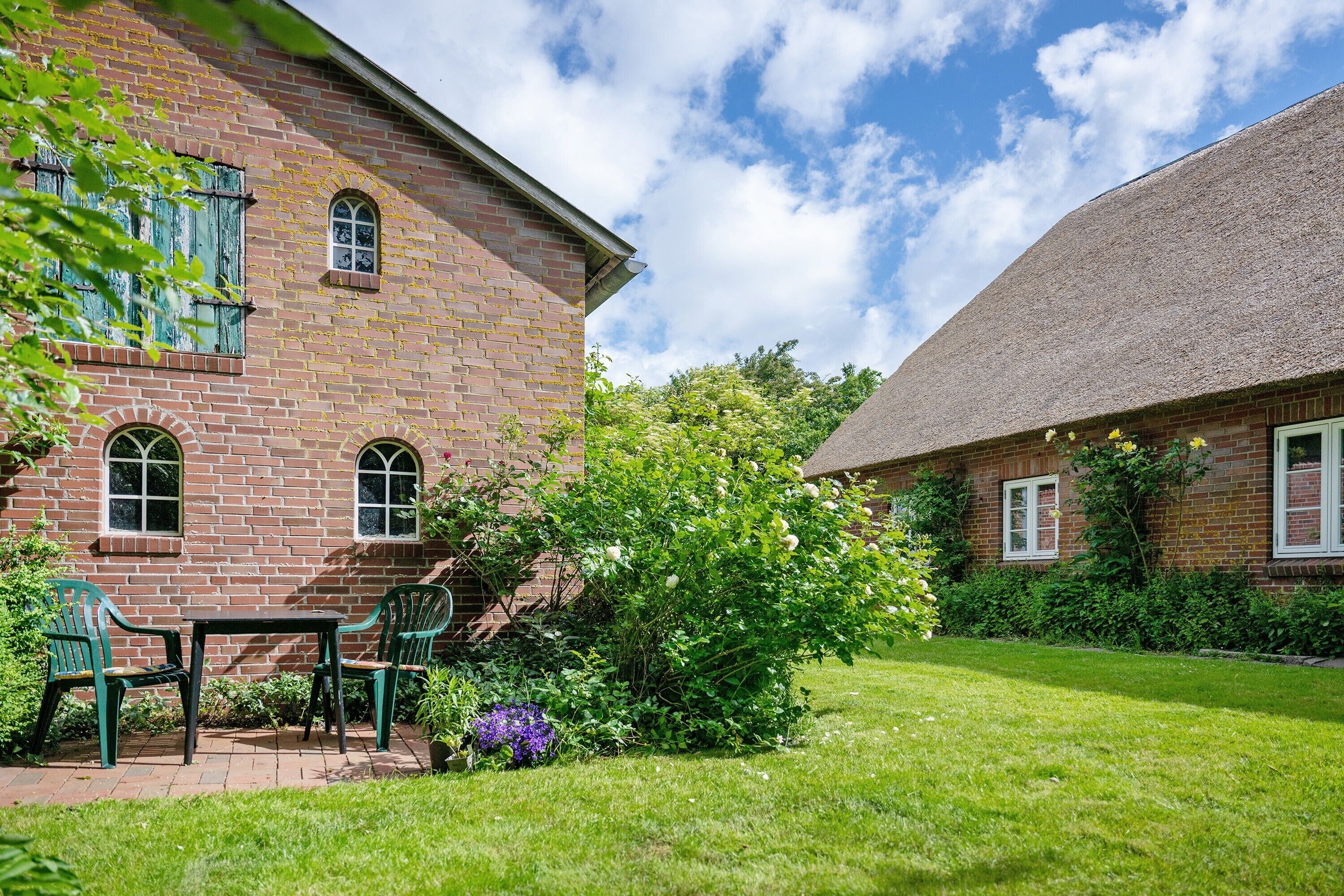 Ein Backsteinhaus mit Strohdach, eine gepflasterte Terrasse mit Tisch und Stühlen sowie ein gepflegter Garten.