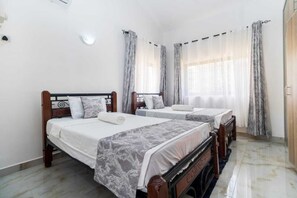 Ein Schlafzimmer mit zwei Einzelbetten, jeweils mit Holz-Kopfteil, weißen Bettwäsche und grauen, gemusterten Bettdecken. Die Wände sind weiß, der Boden ist mit Fliesen belegt und es gibt große Fenster mit durchscheinenden Vorhängen.