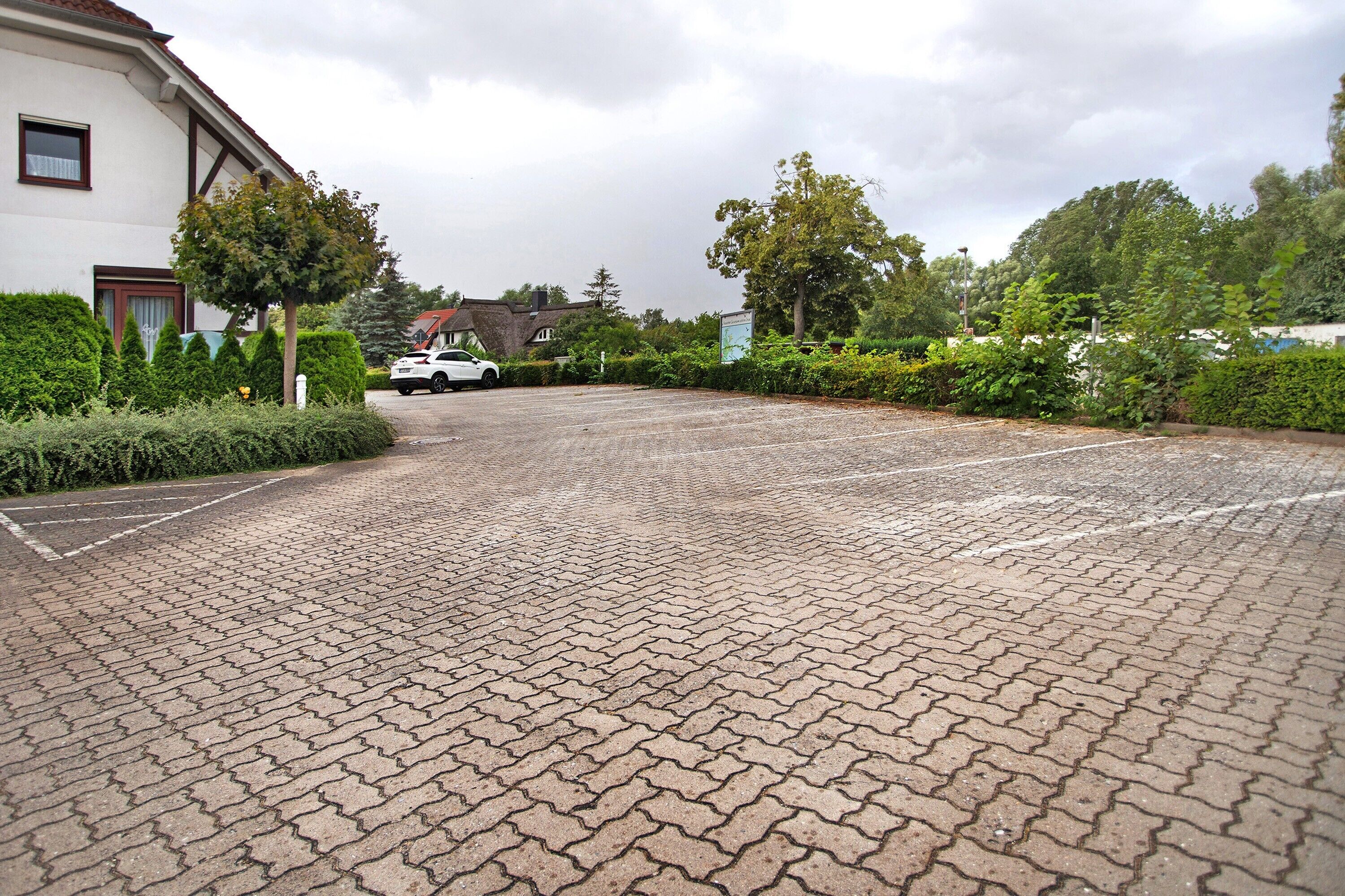 En asfalteret parkeringsplads med en hvid bil parkeret i det fjerne, omgivet af grøn vegetation og boligbygninger.