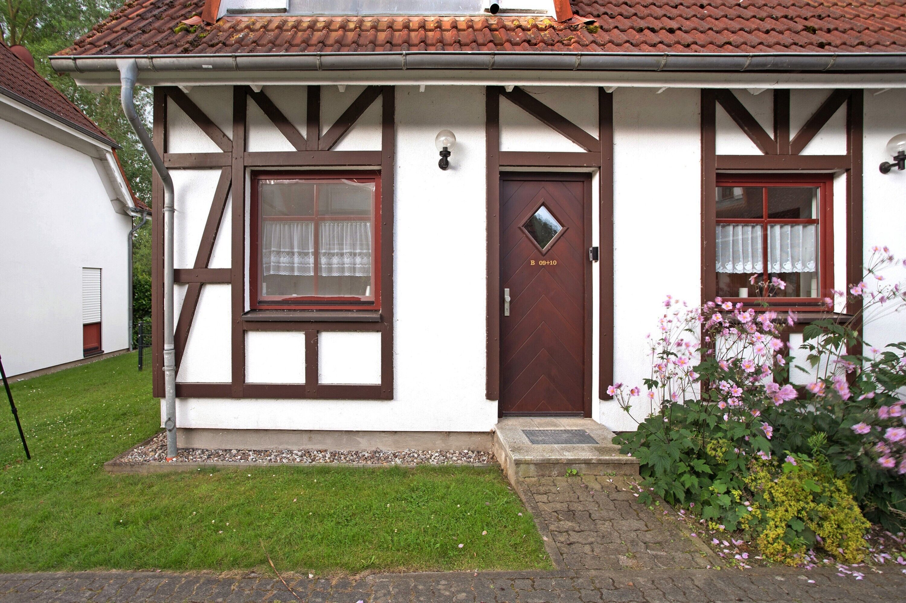 Un bâtiment traditionnel avec une porte marron, deux fenêtres ornées de rideaux et un petit jardin parsemé de fleurs roses.