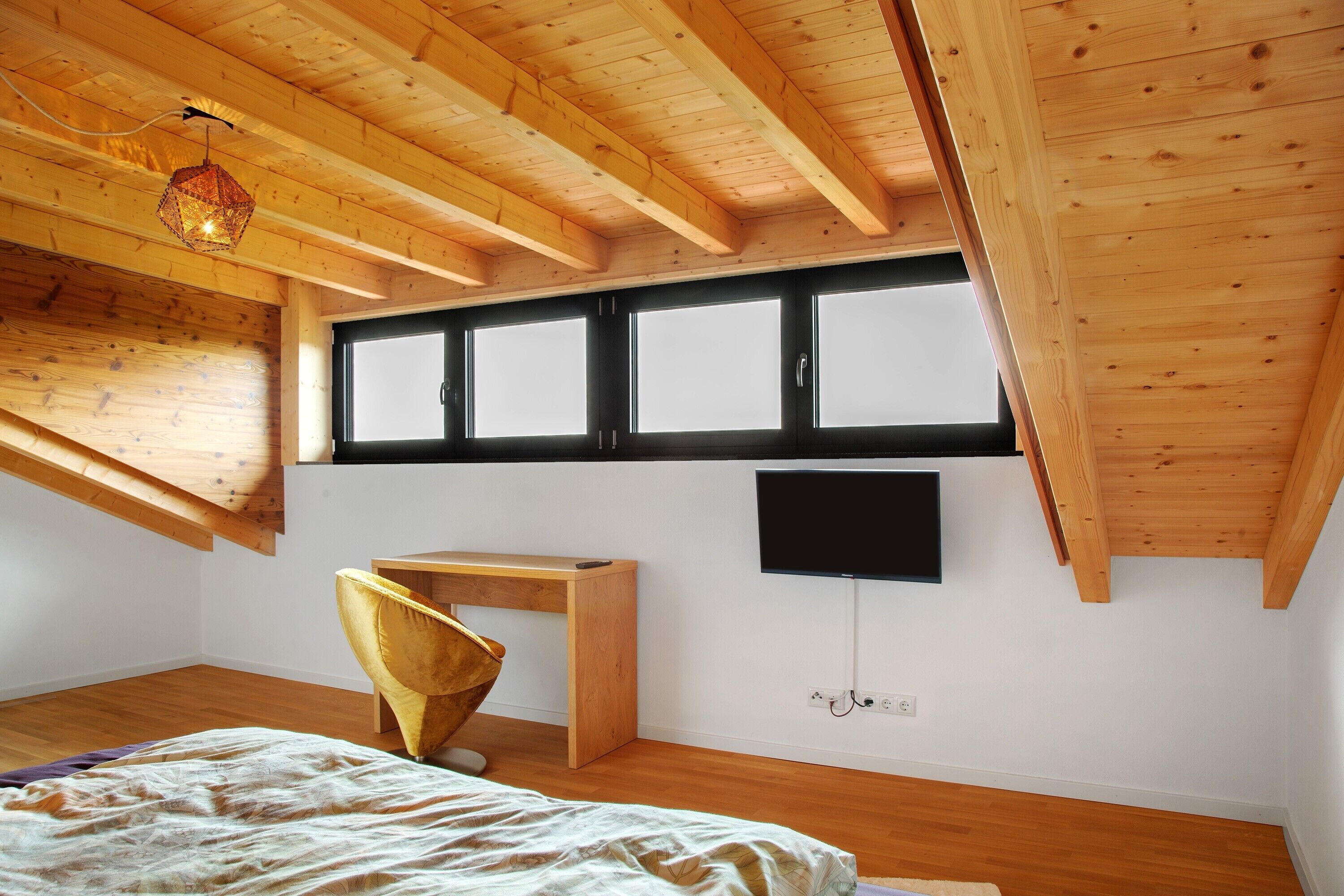 Ein Schlafzimmer mit schrägem Dach, einem Bett, einem Schreibtisch, einem Stuhl und einem an der Wand befestigten Fernseher.