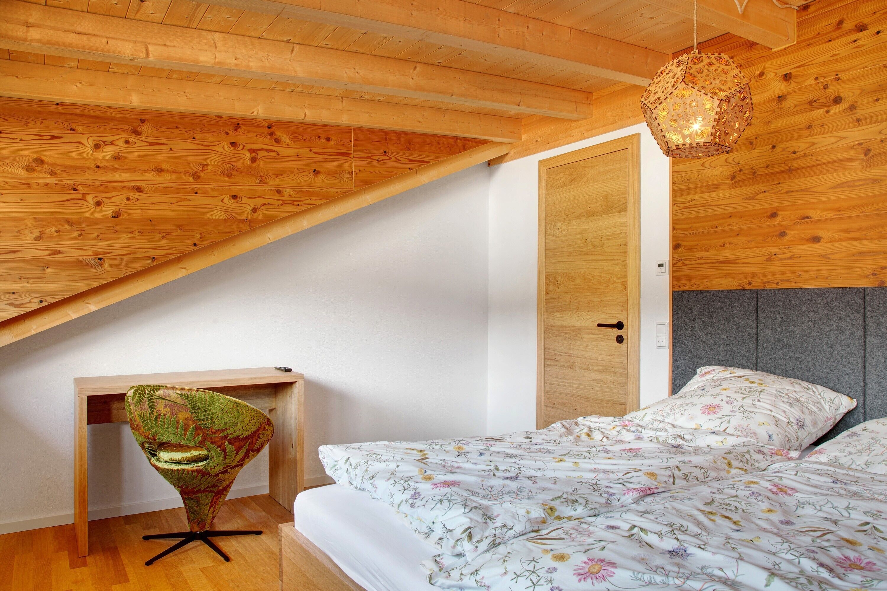 Ein Schlafzimmer mit schrägen Wänden, einem Bett mit floralem Bettbezug, einem Sessel und einer Pendelleuchte.