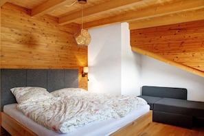 A wooden interior with a bed, a grey headboard, a grey sofa, and a hanging light fixture. - Chalet 'Sonnenwald' with Mountain View, Balcony and Wi-Fi (Grattersdorf)
