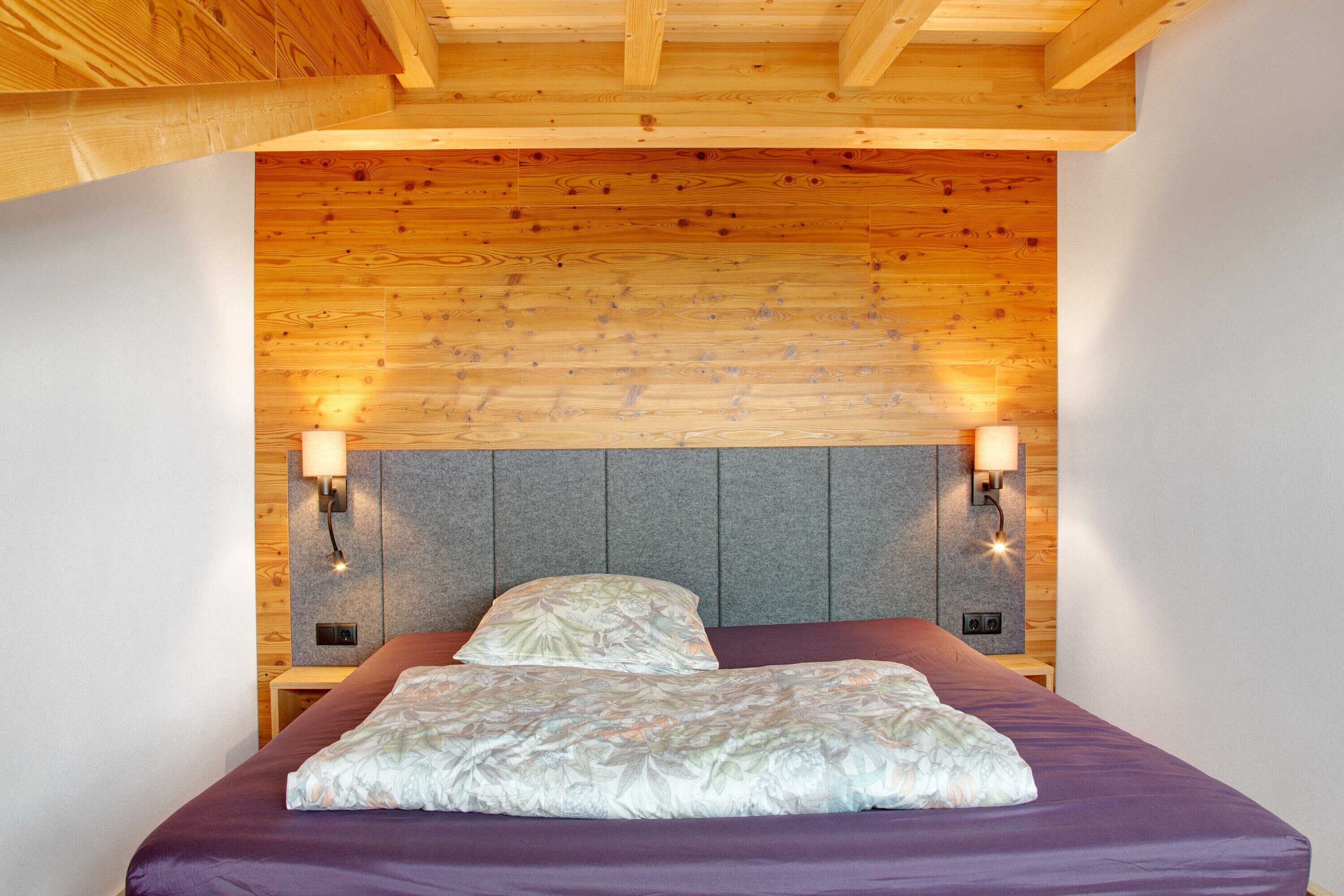 A bedroom with a wooden ceiling, a headboard with wall-mounted lights, and a purple bedspread.