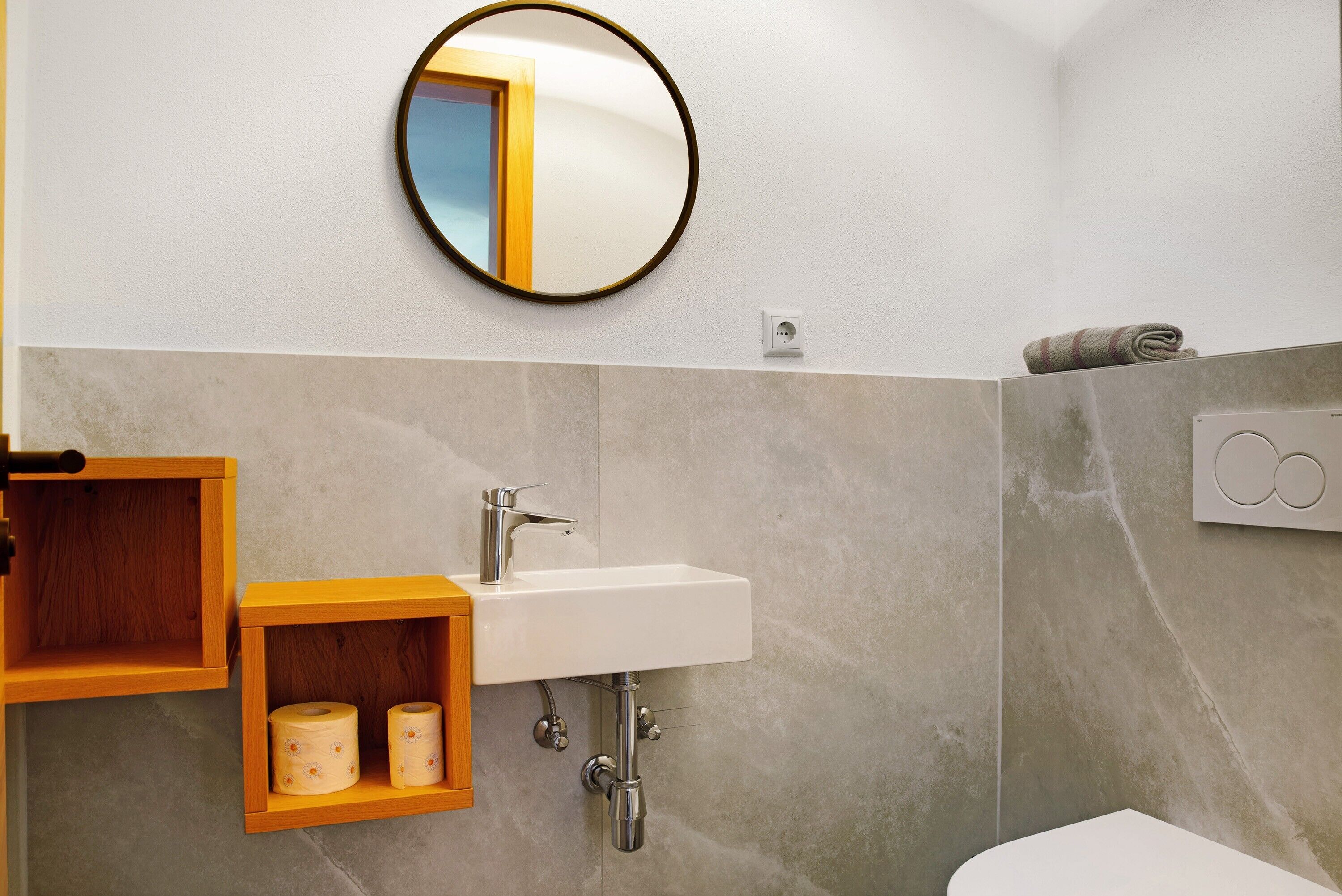Un baño con un lavamanos blanco, un estante de madera con papel higiénico, un espejo redondo y una toalla en la pared.